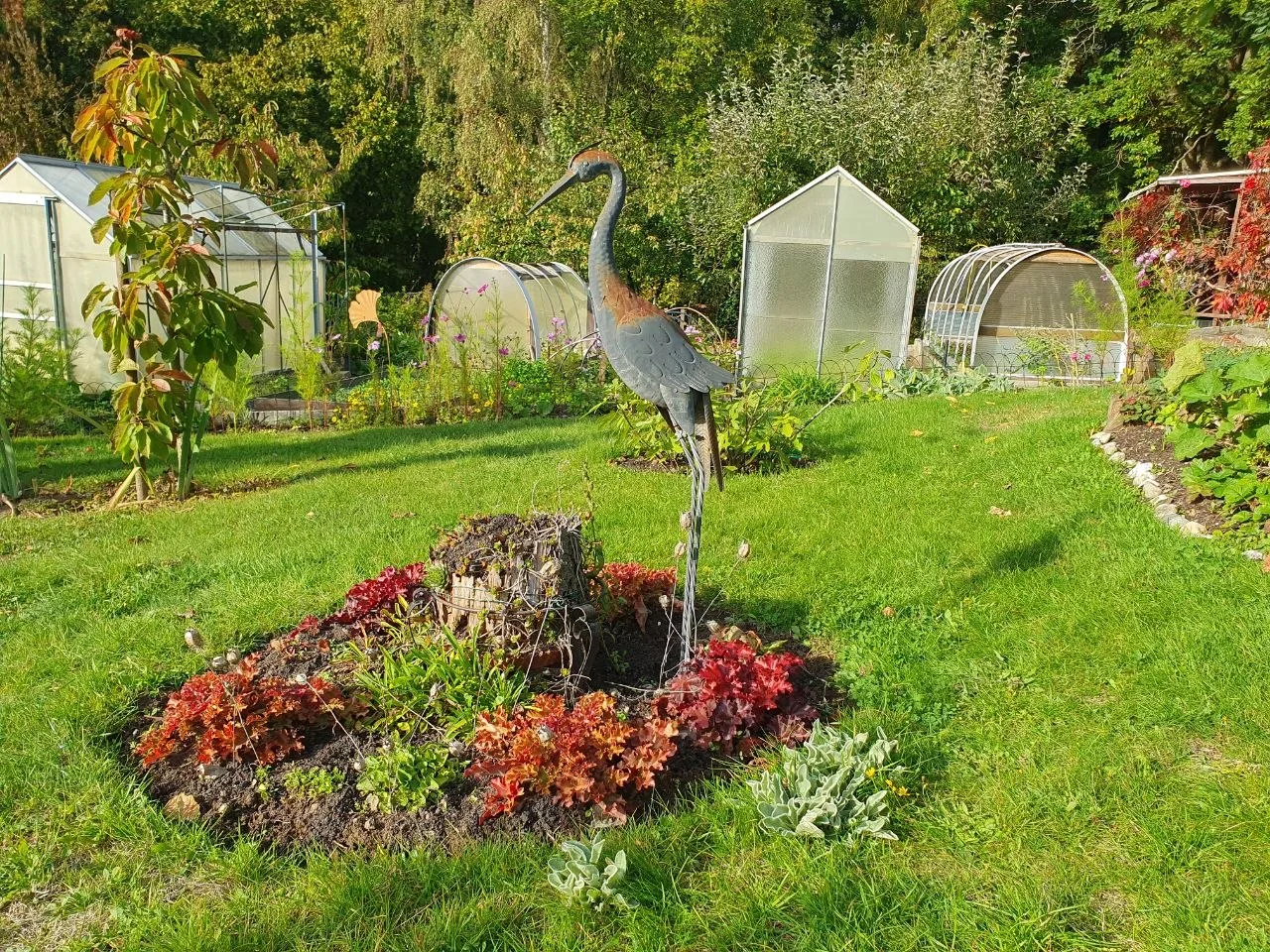 Stilisierte Vogel-Skulptur im Beet, dahinter mehrere Gewächshäuser zwischen Gehölzen